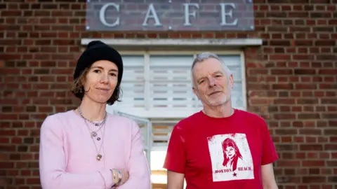 PA Media Emma Fernandez and Patrick Matthews standing in front of the lido cafe. Emma wears a pink cardigan and a black beanie hat, and Patrick wears a red t-shirt.