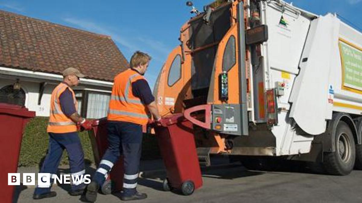 Waste disposal operatives collecting smaller burgundy bins. There are two men wearing orange hi-vis vests loading the bins on to the back of a white bin lorry, with green signage on the driver's side.