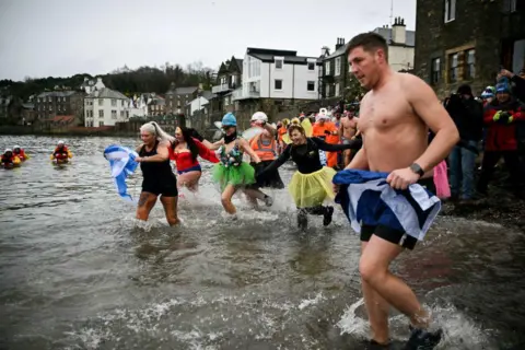 Getty Images Swimmers in colourful outfits running into the water. There are houses in the background on the shore.