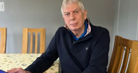 Mike Benbow, a man with white hair and wearing a navy jumper, sits at a kitchen table holding a tablet.