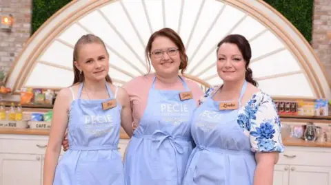 STVR Three women stand side by side in a bright kitchen set, each wearing a light blue apron with a name tag. Behind them are white cabinets, baking equipment, and a decorative arch with a sunburst design.