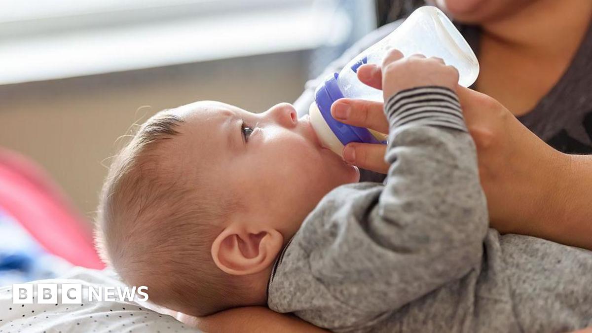 A baby dressed in a grey sleepsuit being held in a woman's lap. The baby is holding the woman's index finger while drinking from a bottle.