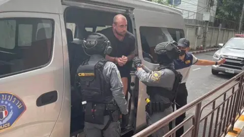 Essex Police Police officers dressed with heavy body armour and helmets lead a handcuffed Ransome out the side of a grey police van.