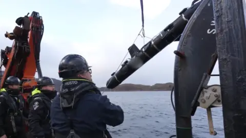 BBC Three men in hardhats onboard a ship launching  a SG-1 Fathom glider into the sea. It is lifted by a pulley and lowered into the water