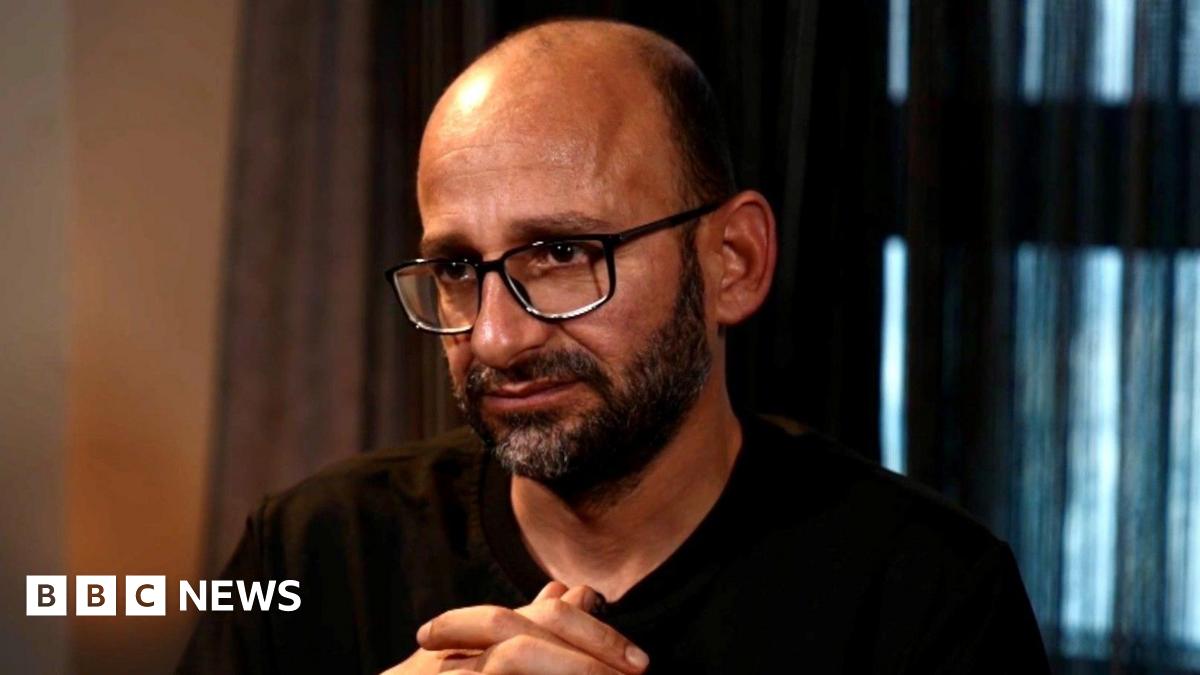 Sami al-Saei during his interview with the BBC. He has a bald head with a short black beard. He wears black-rimmed glasses and a black T-shirt.