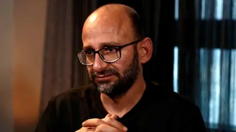 BBC Sami al-Saei during his interview with the BBC. He has a bald head with a short black beard. He wears black-rimmed glasses and a black T-shirt. 