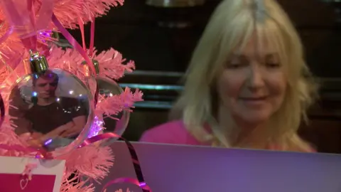 BBC Lisa Wilson smiles at her computer. In the foreground a bauble of her son Tom hangs off a pink Christmas tree.