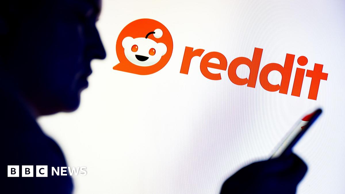 In this photo illustration, a silhouetted individual is seen holding a mobile phone with a Reddit logo displayed in the background