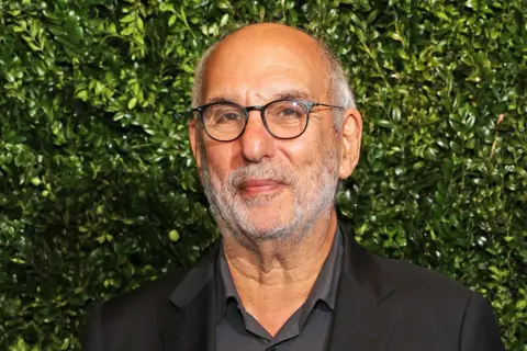 Alan Yentob is wearing a black collared shirt layered with a black blazer. The background consists of a dense green wall of foliage, creating a natural and vibrant backdrop. The lighting is even, highlighting the texture of the greenery and the dark clothing.