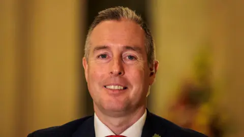 PA Media Paul Givan inside Stormont, he is wearing a navy suit and a white shirt with a red tie. He has short white hair.