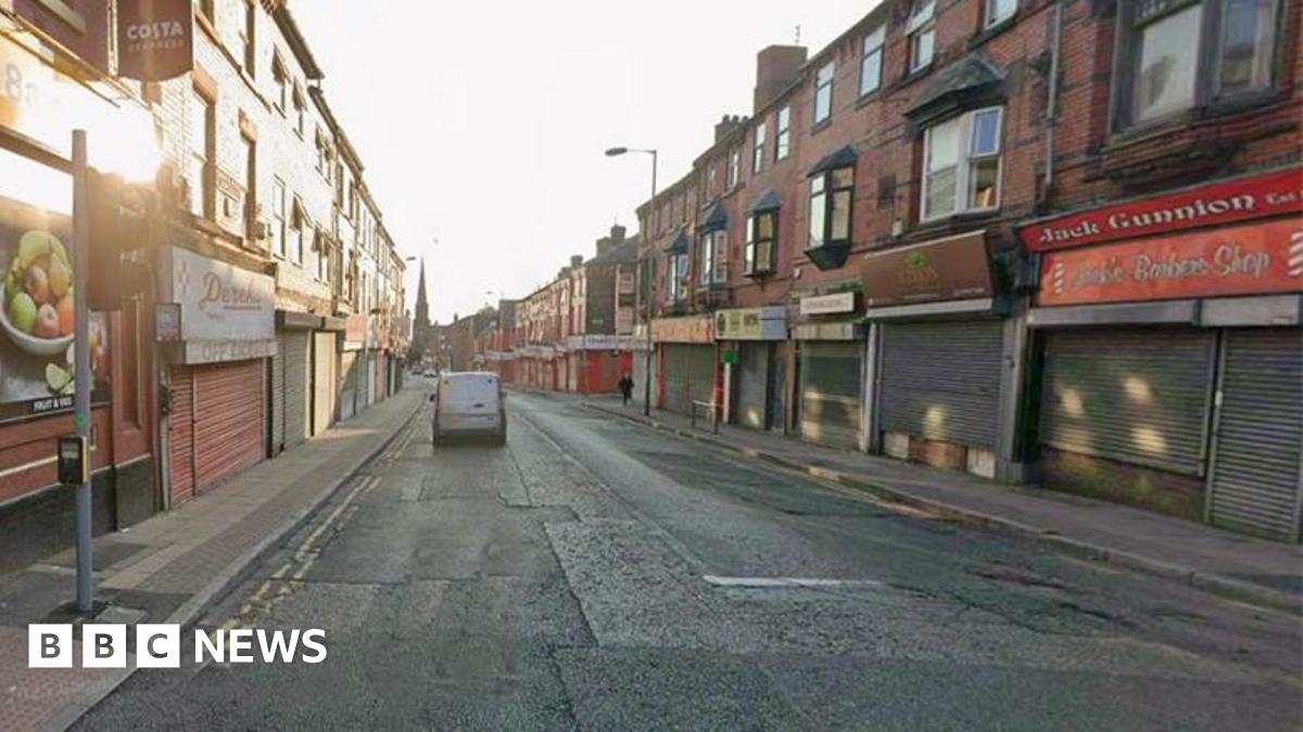 A white van travelling down Breck Road on a clear day. There are a number of shops with shutters down lining the road.