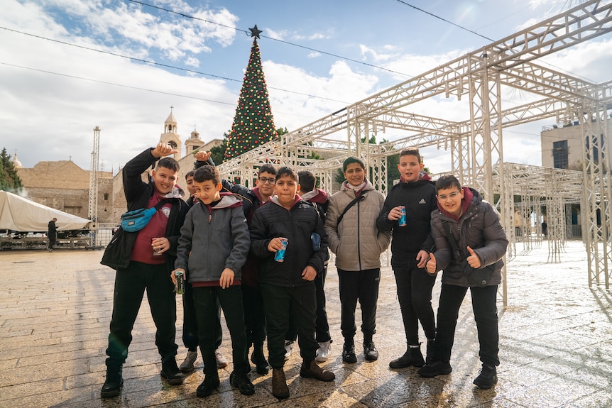 A group of children standing together, making gestures and poses, in the distance behind them is the top of a christmas tree.