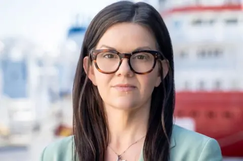 Kirstie Langan Kirstie Langan looking at camera, she is wearing a light mint coloured jacket and there are offshore vessels in the background in Aberdeen.