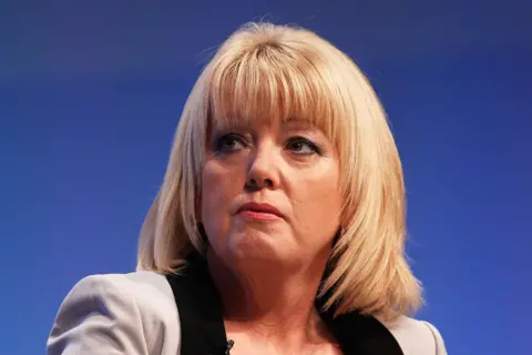 Getty Images Baroness Newlove is wearing a light gray blazer with black lapels over a black top. Her hairstyle is straight and shoulder-length, with a side part. The background is a solid blue colour, suggesting a formal event or conference setting.