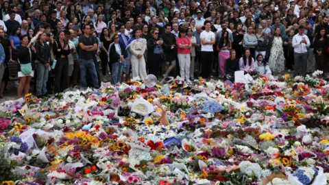 Reuters People stand next to floral tributes for the victims of a shooting at Bondi Beach in Australia.