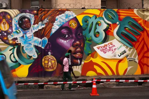 Sodiq Adelakun / REUTERS People walk past a brightly coloured mural featuring an astronaut and a woman with a purple stripe over her face, along with African artefacts and a message saying: Rewrite your own story