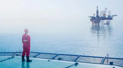 Getty Images An oil worker in a red uniform and and hard hat stands on one North Sea platform looking across the water to another platform.