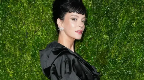 Getty Images Lily looking at the camera over her shoulder in a black coat standing in front of a green  background