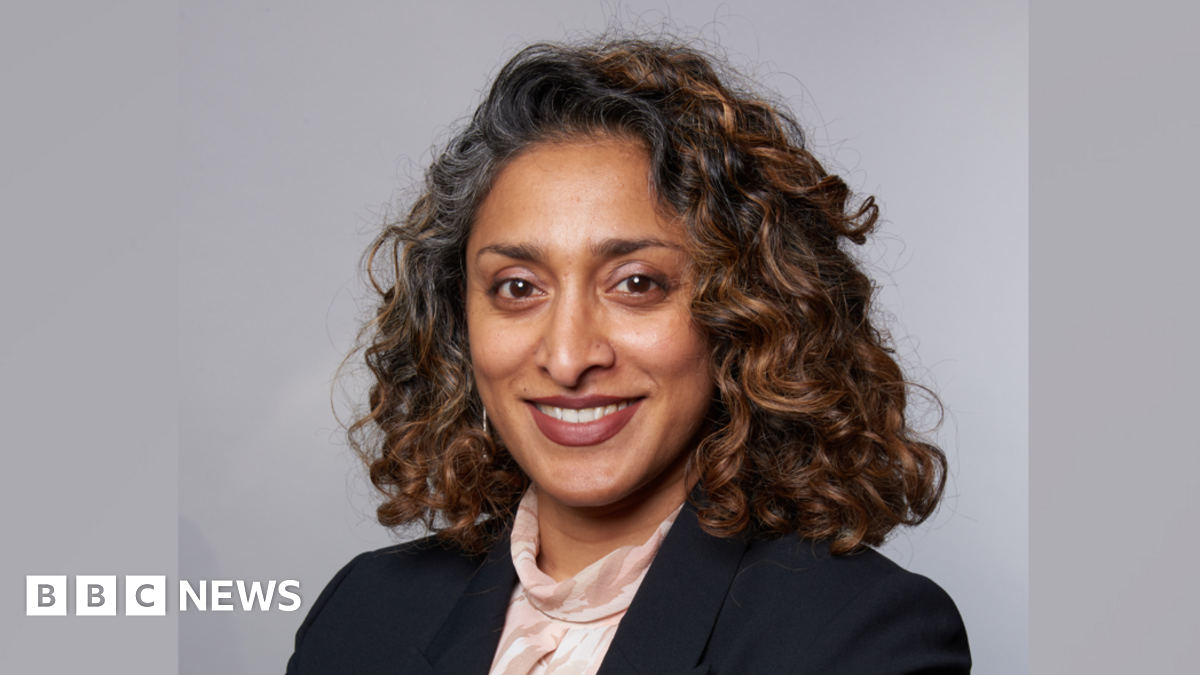 A head and shoulders image of Monisha Gower. She is wearing a black blazer over a light top. She is standing in front of a grey backdrop and smiling while looking into the camera.