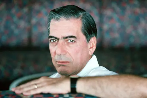 Getty Images Mario Vargas Llosa is seated indoors, wearing a white collared shirt. One arm is resting on a surface, showing a black wristwatch and a gold ring on the hand. The background features a patterned fabric with a mix of muted colours, creating a soft, textured backdrop. 