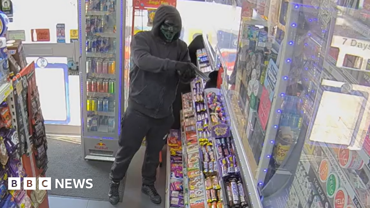 A man wearing a black hooded jacket, black trousers, black shoes, black gloves and a black horror-mask standi ng in a shop pointing what appears to be a black pistol. He is standing inbetween a rack filled with bags of sweets and a shelf filled with bars of chocolate.