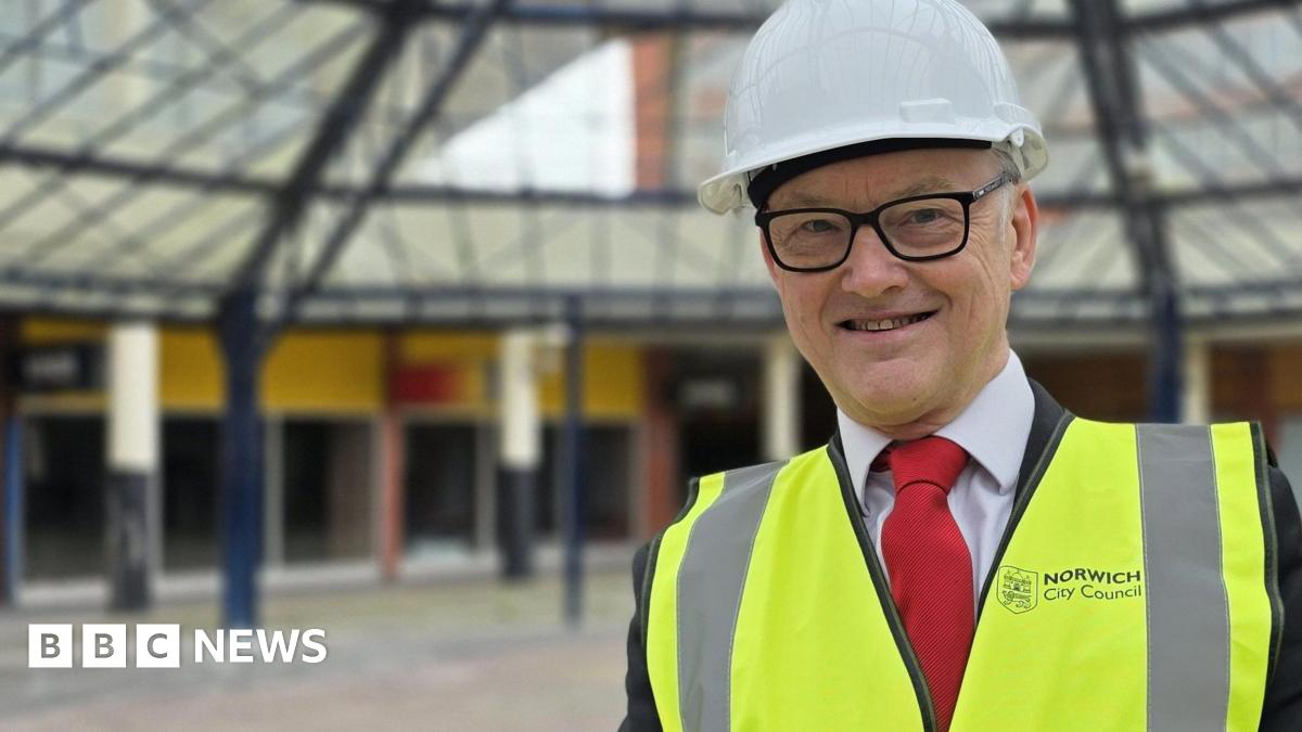 Mike Stonard is wearing a high-vis jacket over a suit with a red tie. He has a white hard hat on his head and is standing beneath a glass canopy.