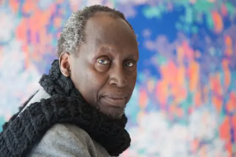 Getty Images Ngugi Wa Thiong'o is wearing a thick, chunky black knitted scarf draped around the neck and a light gray coat. The background features a vibrant, abstract artwork with splashes of blue, pink, orange, and green colours, creating a lively and artistic setting. 