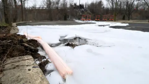 Bastiaan Slabbers/Getty Images White firefighting foam spills out across a concrete ground, it is contained with a orange bund. In the background is a chain link fence with trees surrounding and orange bollards. 