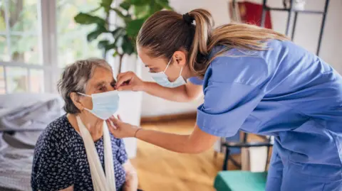 Getty Images A nurse wearing scrubs and with a face mask on checks an elderly woman, also wearing a face mask, in a nursing home