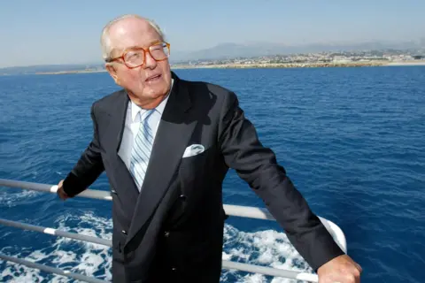 Getty Images Jean-Marie Le Pen wearing a dark navy suit with a light blue tie and white pocket square stands on the deck of a boat, holding onto the railing. The boat is on a calm blue sea.