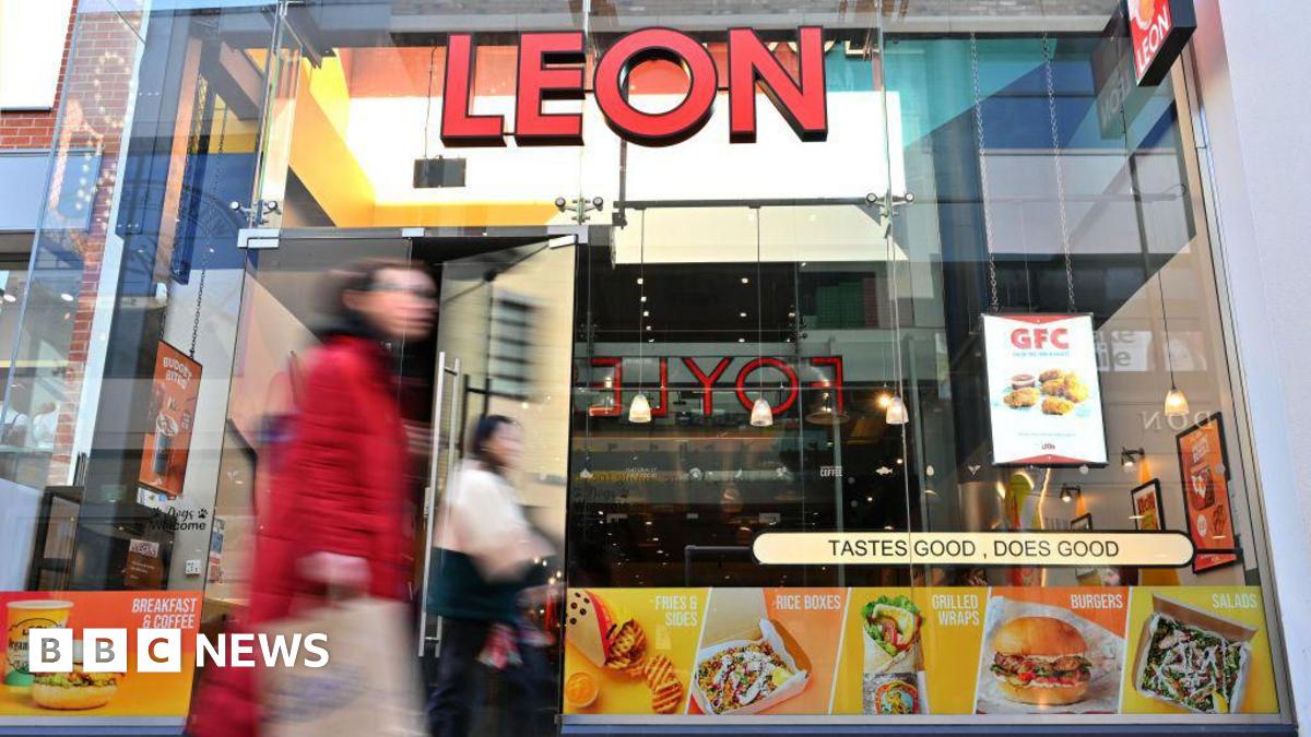 A blurry woman in a red jacket walks past a glass store front with pictures of fast food and a red "LEON" sign.