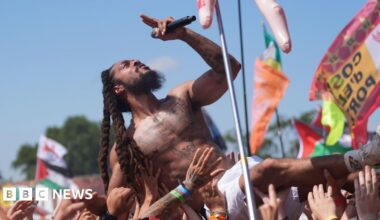 Bob Vylan crowd surfs during his performance at Glastonbury Festival. He is topless and is wearing white shorts and holding the mic in his left hand