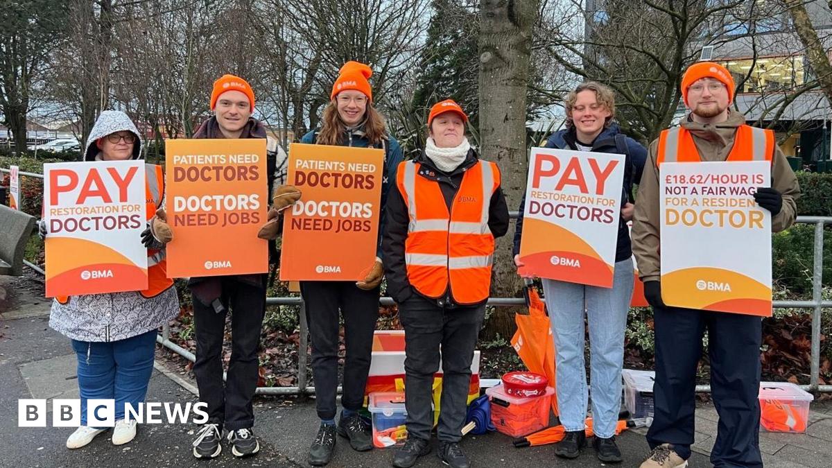 Five people standing outside Hull Royal Infirmary wearing orange beanie hats and holding placards. One reads: "Pay restoration for doctors". Another says: "Patients need doctors, doctors need jobs".