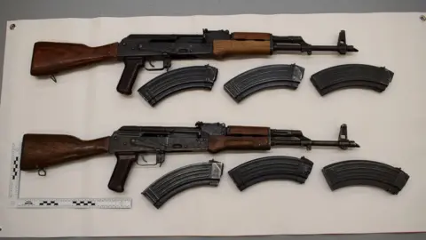 Greater Manchester Police Two rifles surround magazines on a white background.