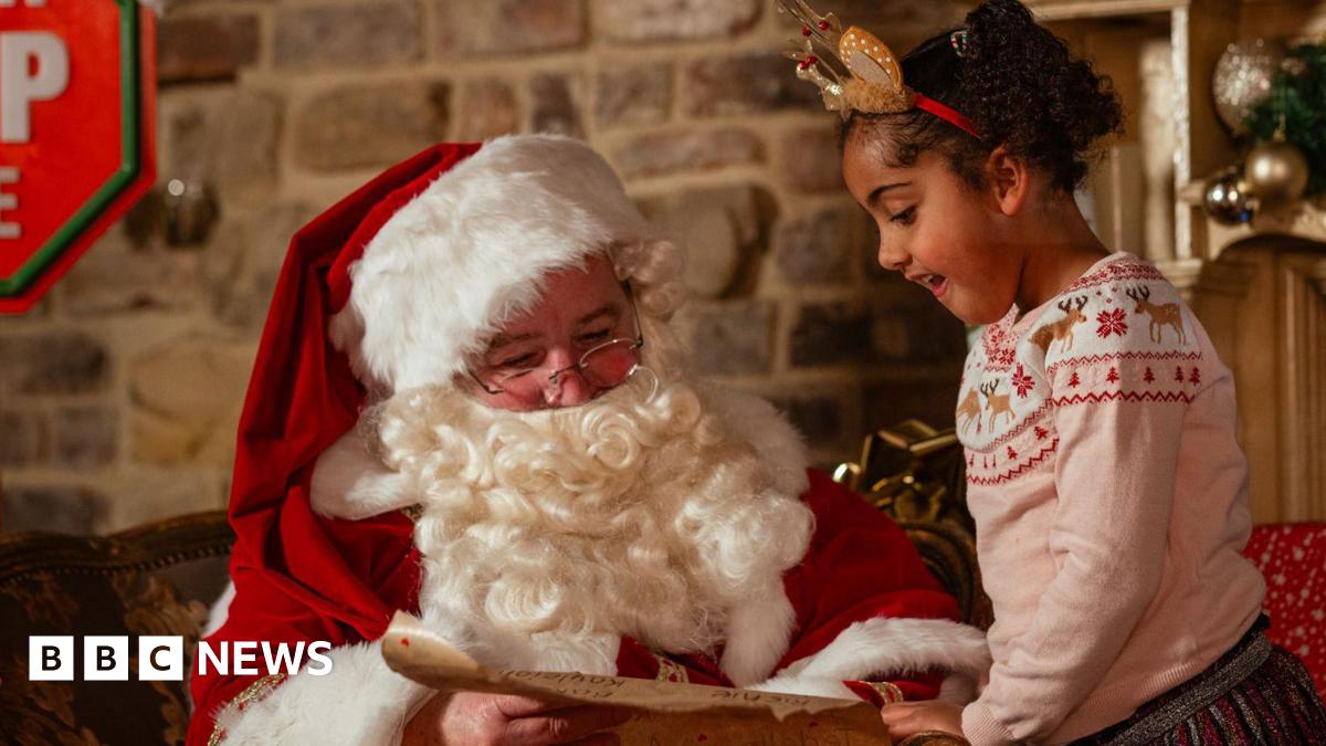 A man dressed as Father Christmas in traditional white-trimmed red clothes is leaning over a book, while a child of around eight years old, wearing a Christmas decoration headband stands by him also leaning over the book.