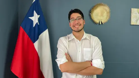 Jeremías Alonso poses for a photo. He is standing next to a Chilean flag, wearing a white shirt with its sleeves rolled up. He has crossed his arms and is smiling at the camera. 