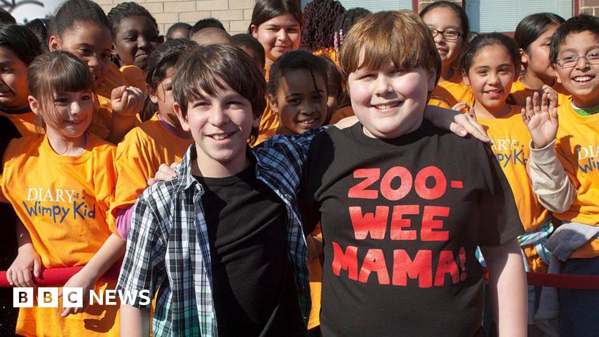 Two children stand with their arms around each others' shoulders. Behind them are many more children wearing orange t-shirts with the "Diary of a Wimpy Kid" logo.