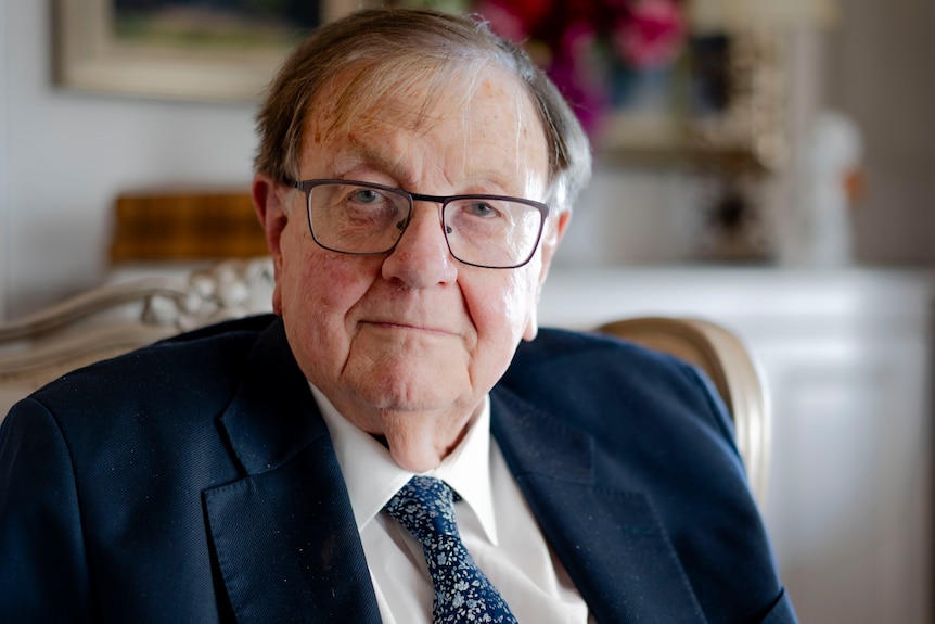 Anthony in a suit, glasses, smiles sternly, he is older, receding hair, sits in a nice ornate chair, painting behind blurred.