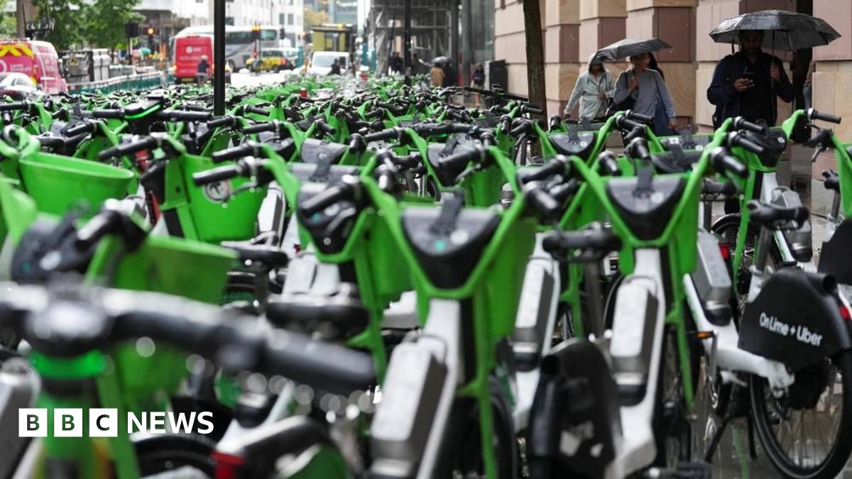 A pavement and road is filled with hundreds of parked, bright green Lime e-bikes. A few people with umbrellas are walking on the side.