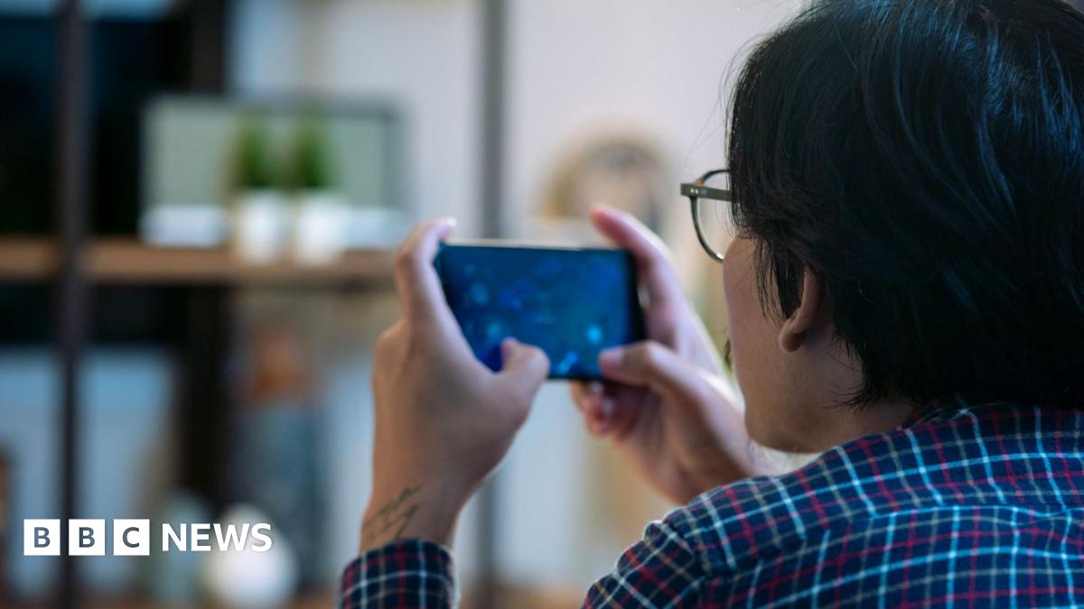Back of the head of a man wearing glasses sitting down playing a video game on a mobile phone