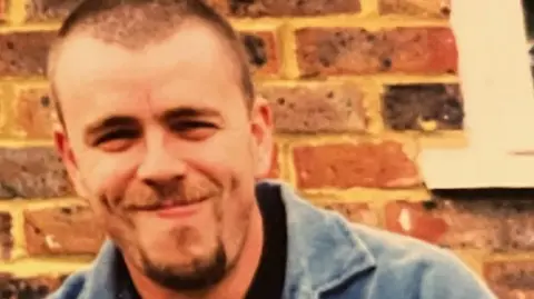 Alison Mark Jenner, pictured by Alison, in front of a red brick house. He wears a denim jacket. 