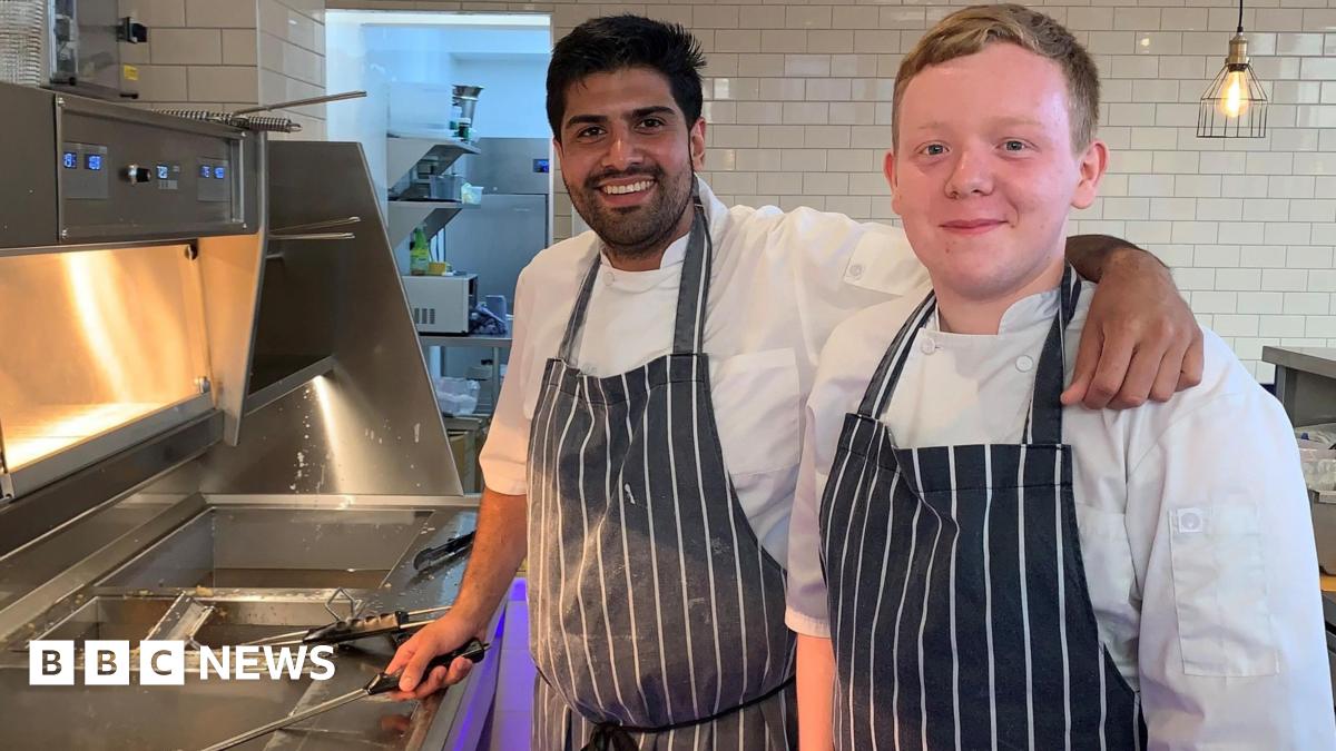 Raj Khaira on the left with one of his colleagues behind the fish and chip shop counter, wearing a white and blue stripped apron.