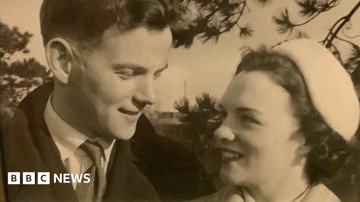 A black and white photo of a young man and woman looking at each other affectionately. The man is wearing an overcoat, a light shirt and a striped tie. The woman has short, curly dark hair under a light hat and is wearing lipstick.