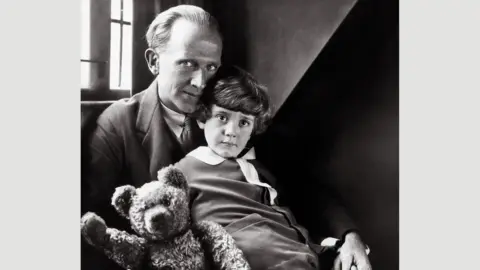 Pictorial Press Ltd/Alamy Black and white image of AA Milne in a suit with his young son Christopher Robin on his lap holding a toy bear in 1926 