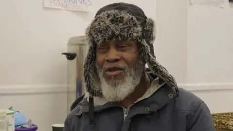 Elijah Augustus in a dark coat and hat with a fur trim. Behind him is a metal container for hot drinks. 