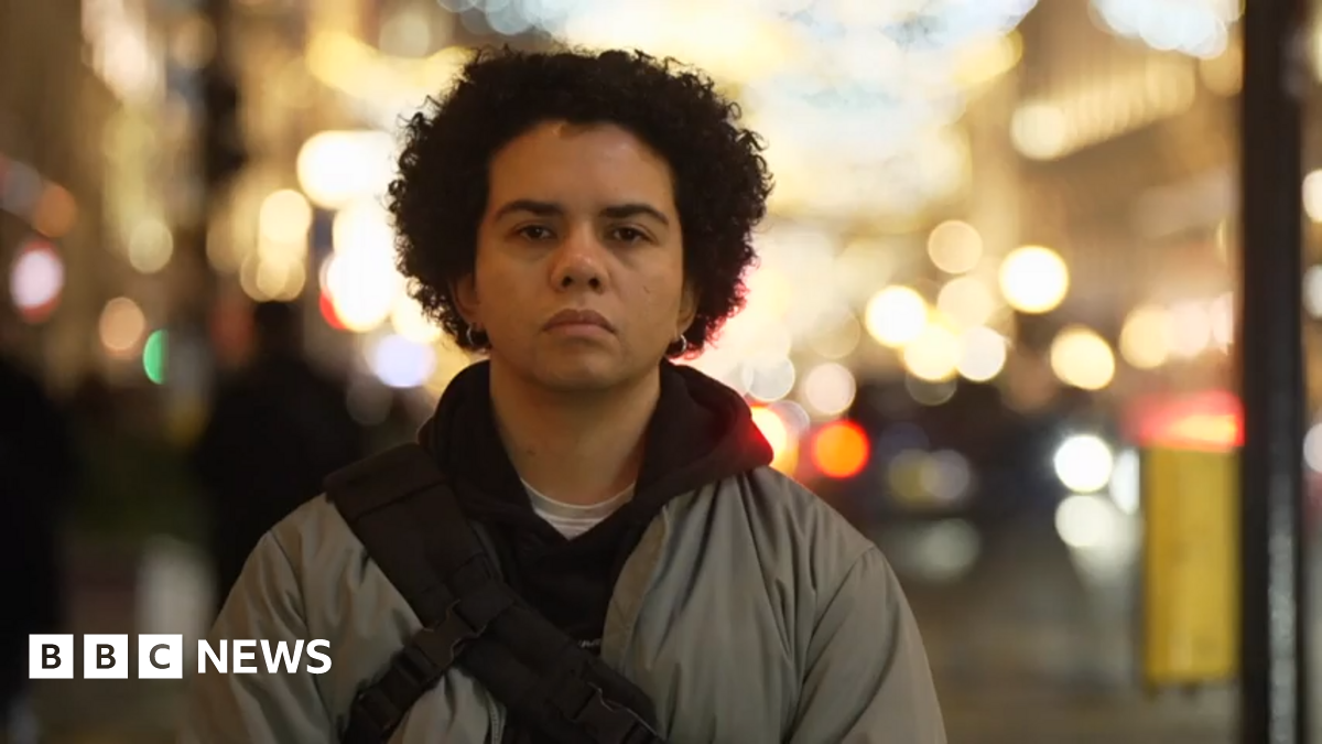 Keira Bell stands in front of a dark, black curtain, wearing a black hoodie with red and yellow designs near the elbows. She has dark, curly, short hair, dark brown eyes and wears earrings in her ears.