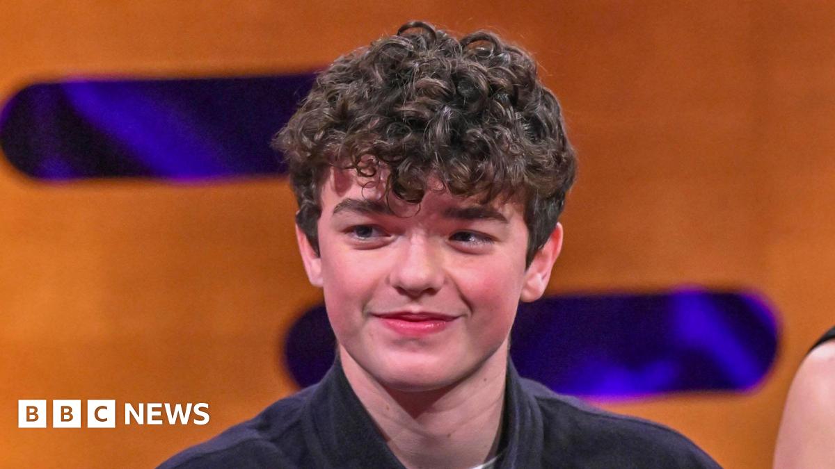 Owen Cooper smiles during Graham Norton interview. He has a mop of dark curly hair and wears a dark shirt. The backdrop is an orange wall decorated with purple lozenge shapes. It is a head shot of him.
