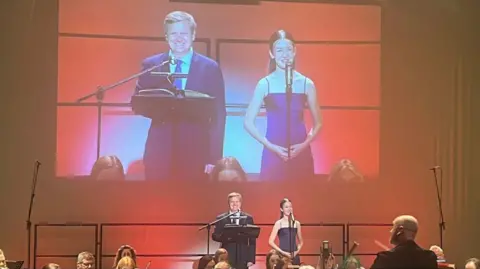 Handout Lorretta, a young girl in a navy strappy dress and Aled Jones, a man in a navy suit, standing on stage behind microphones smiling. Above them is a large projection of the singers.