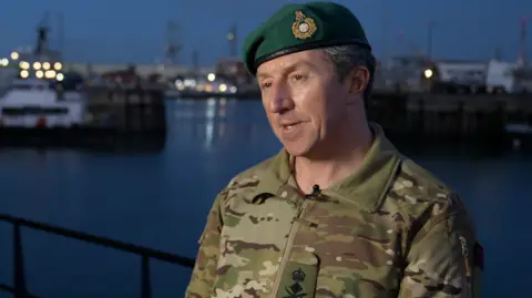 First Sea Lord, General Sir Gwyn Jenkins is in military fatigues wearing a green beret. Behind him is a harbour at dusk.  