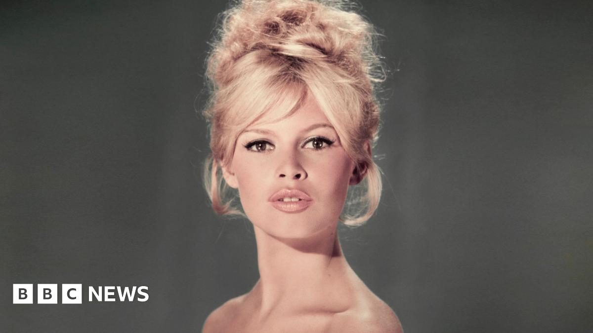 A studio portrait of Brigitte Bardot wearing a light blue off-the-shoulder dress with a blonde beehive hairdo and her lips slightly parted in a pout.